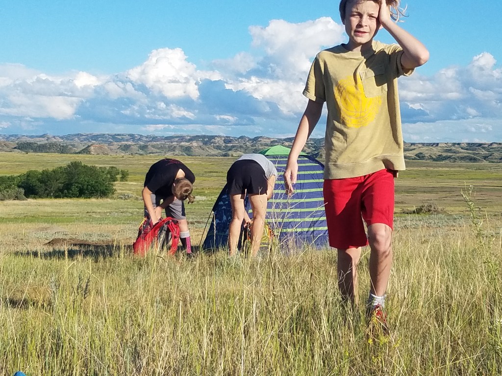 Eliot, Mama Larshop finish setting up the boys' tent, while Quincy comes over to check on dinner's progress.
